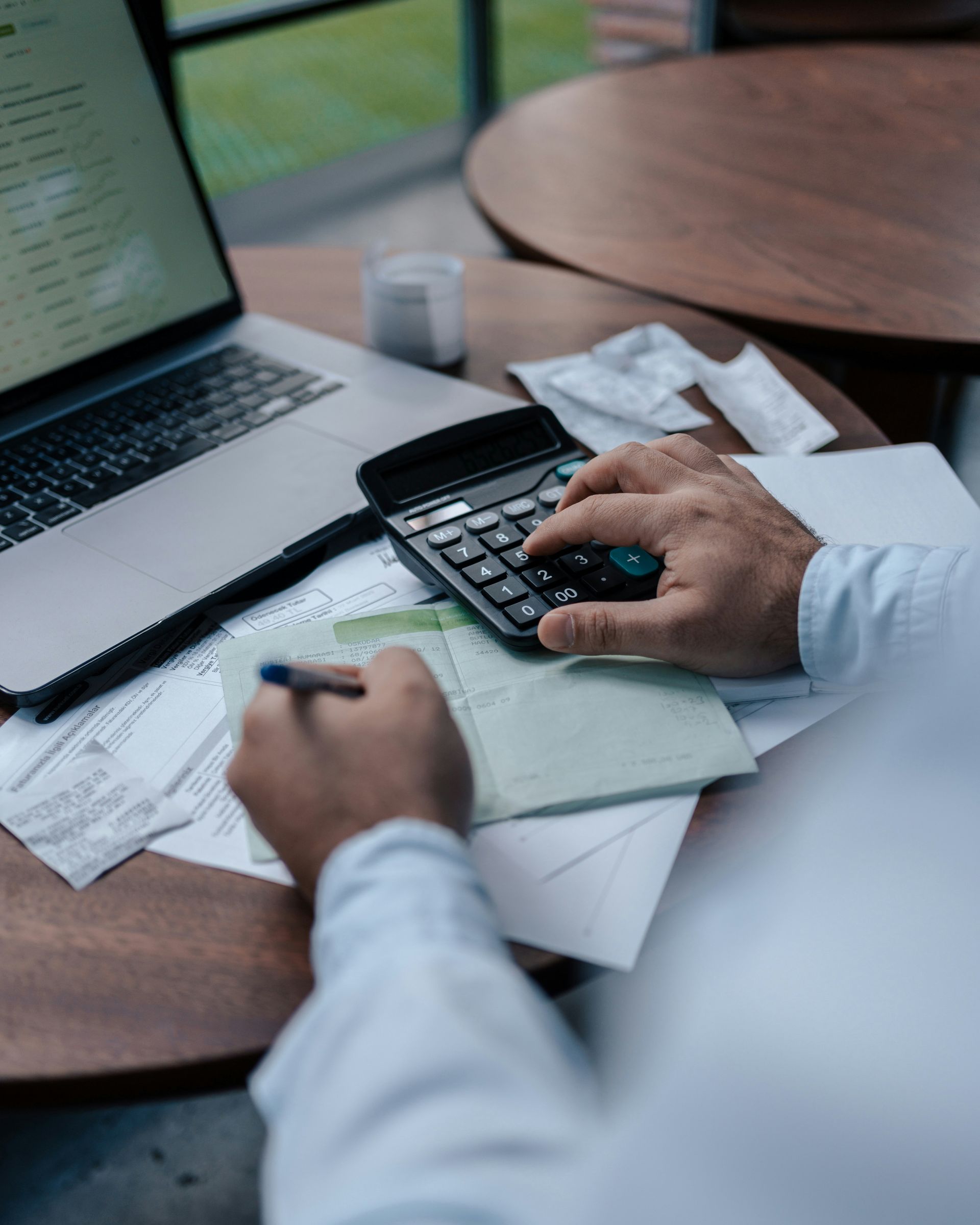 Person using calculator and writing on paper at a table with a laptop, receipts, and a drink.