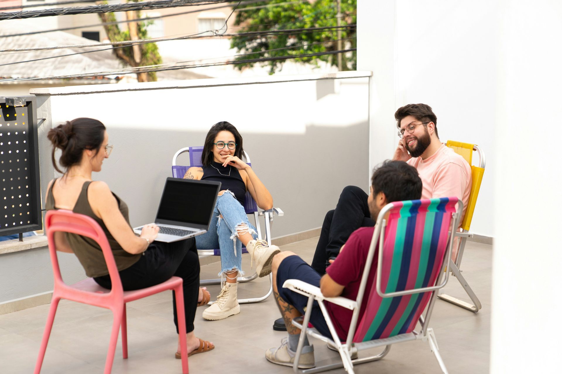 Four people in casual clothes on a patio, sitting in folding chairs, some with laptops and talking.
