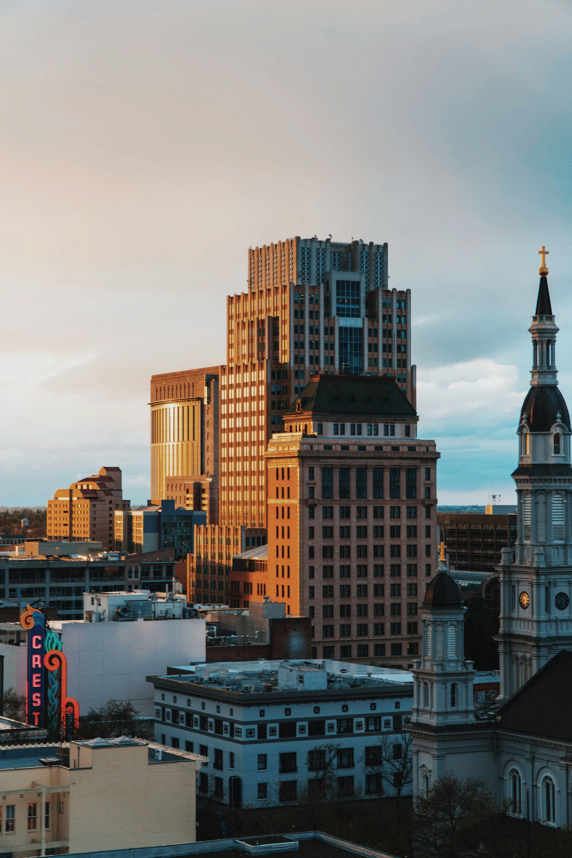 City skyline at sunset; buildings of varying heights with golden light, cloudy sky.