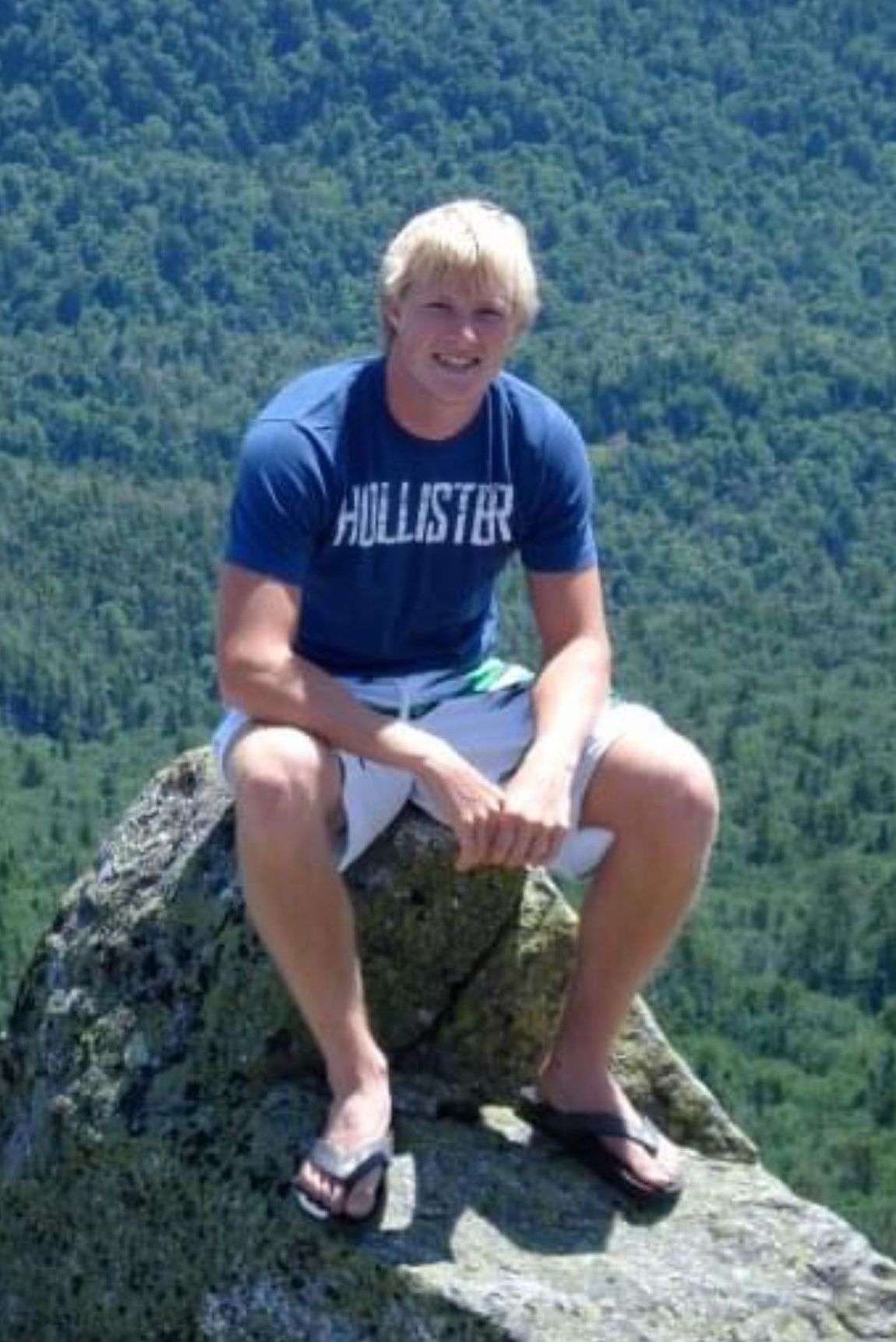 A man wearing a hollister shirt is sitting on a rock.