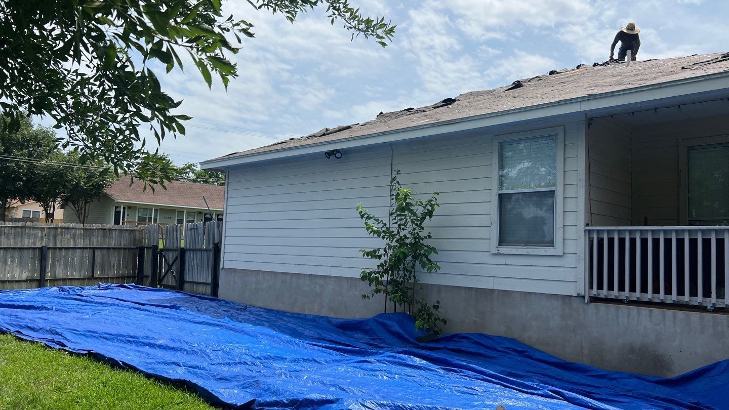 A man is standing on top of a white house with a blue tarp in front of it.