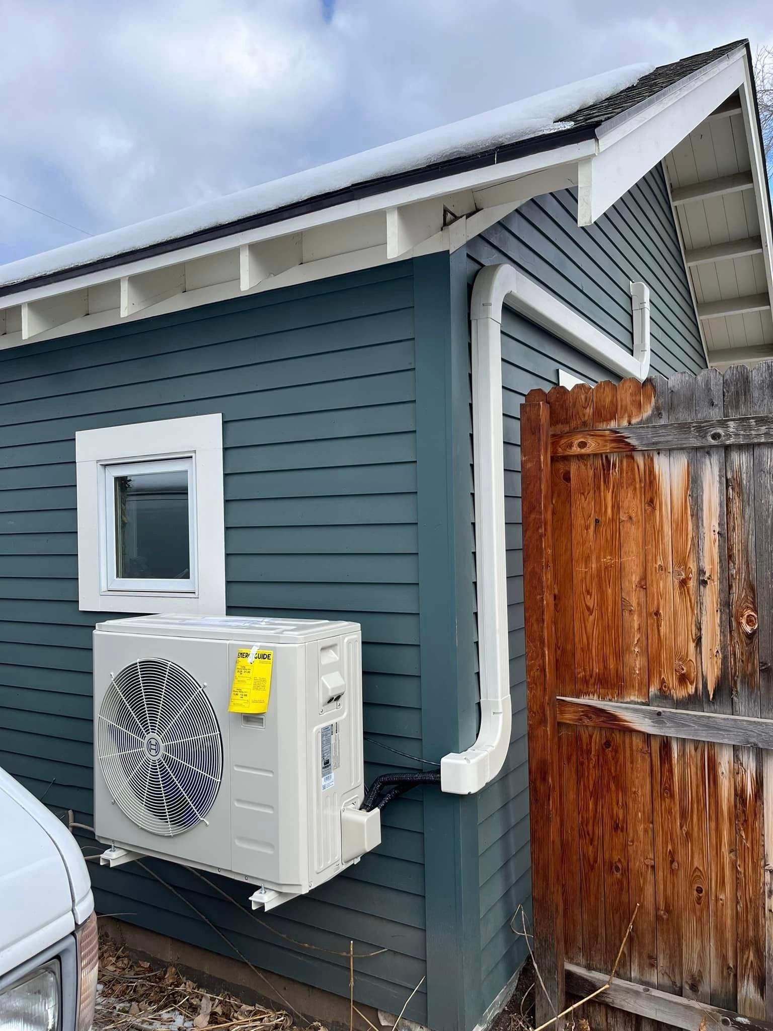 An air conditioner is installed on the side of a house next to a wooden fence.