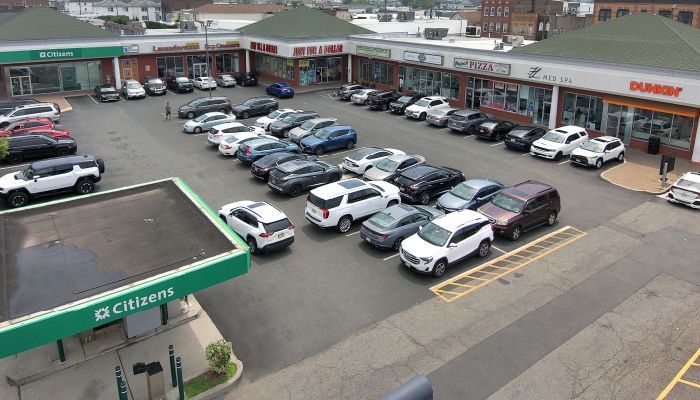 A commercial strip mall with parked cars, a gas station, and various storefronts on a sunny day.