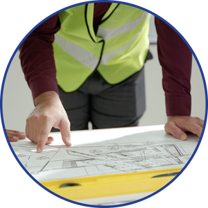 Person in a yellow safety vest points at construction plans laid on a table; a level is in front.