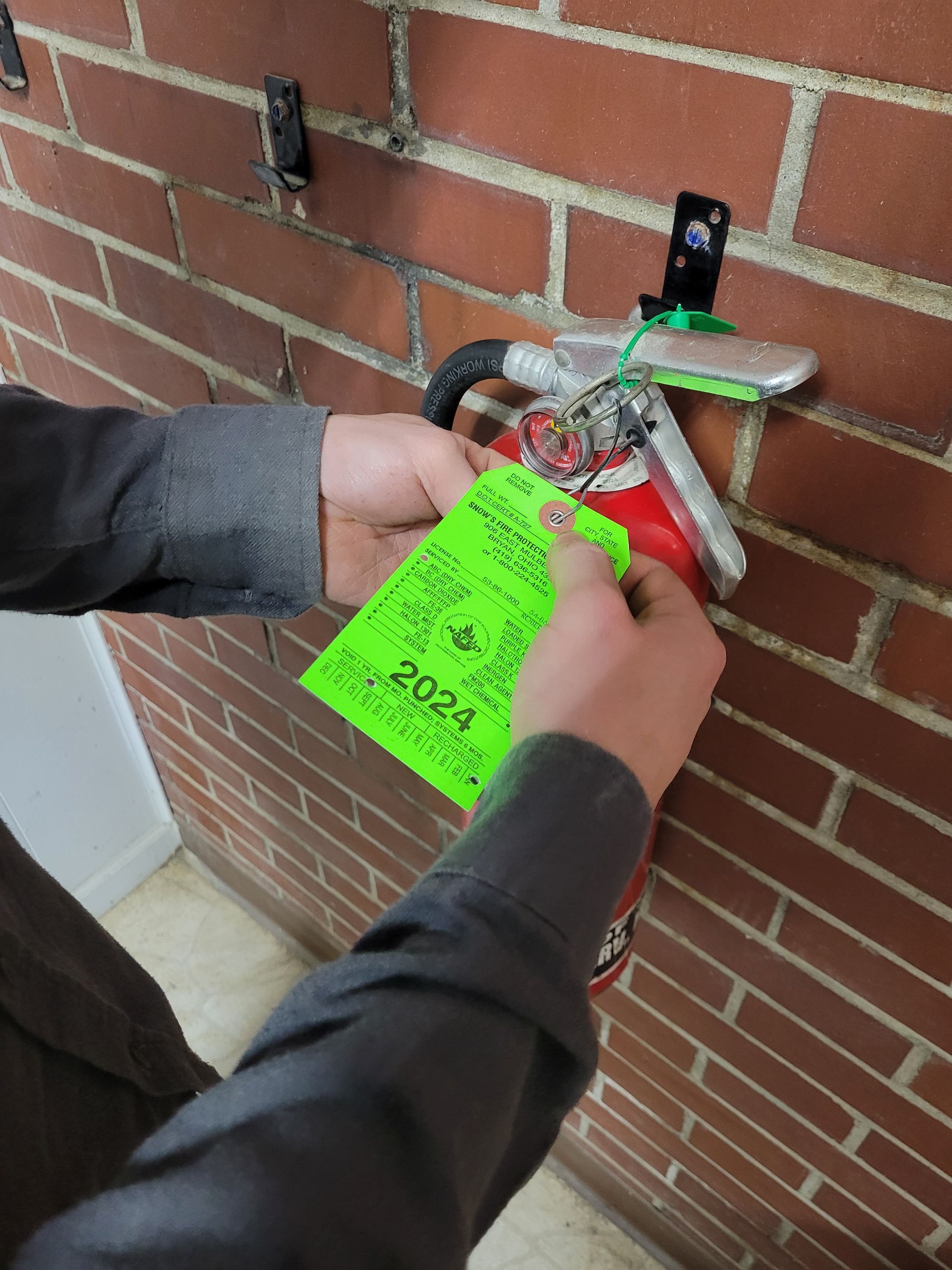Person attaching a green inspection tag to a red fire extinguisher mounted on a brick wall. Tag reads “2024”.