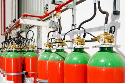 Red and green fire suppression cylinders in a row, connected to red pipes, mounted on a white wall.