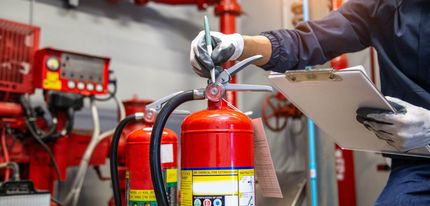 A person inspects a fire extinguisher in an industrial setting. They are wearing gloves and using a pen and clipboard.
