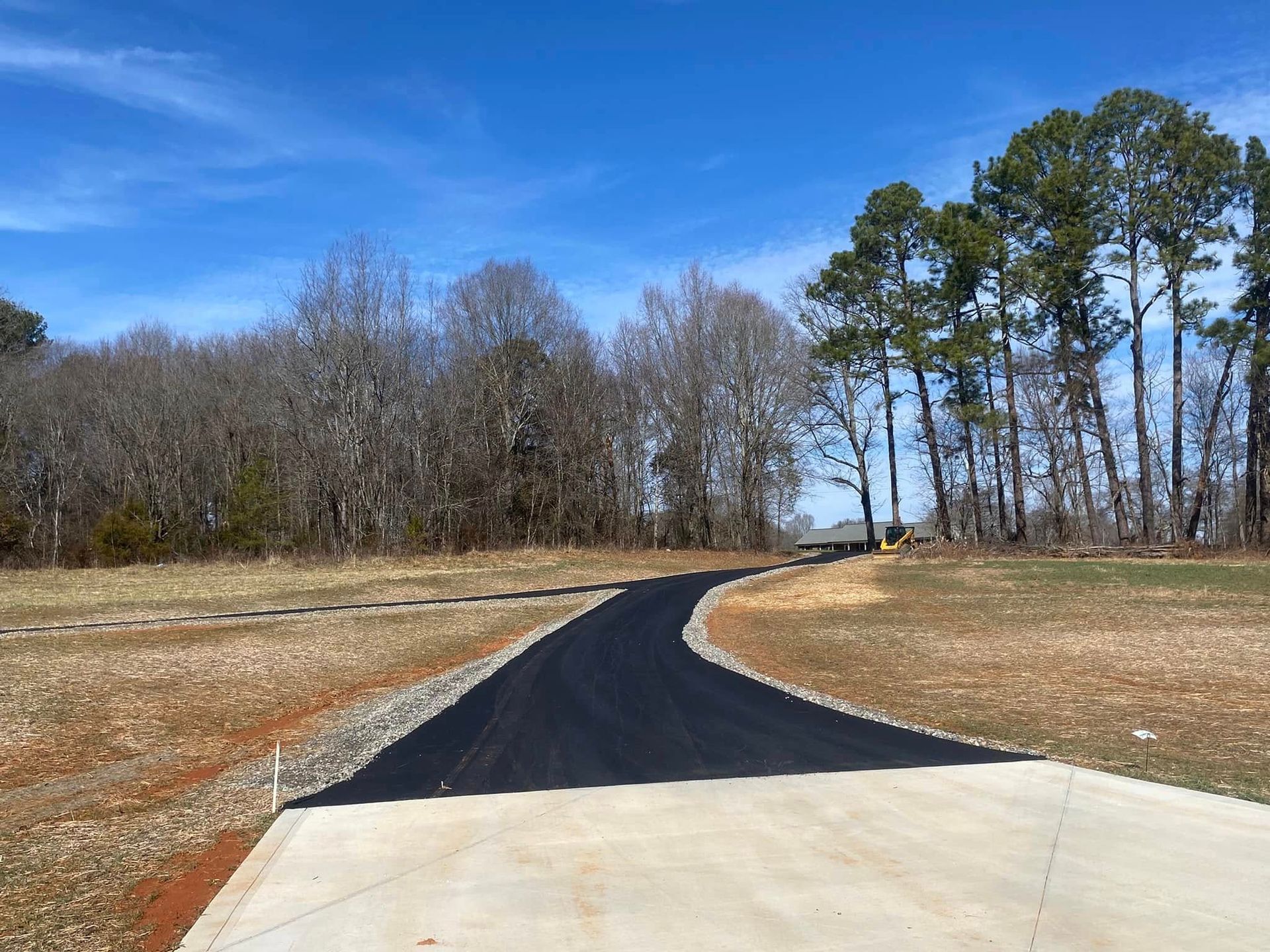 Curved black paved driveway through a brown grassy field with trees under a blue sky