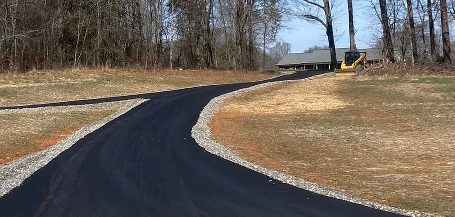 Freshly paved black driveway curving through a grassy, leafless wooded area