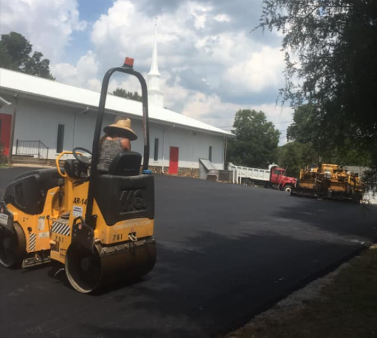 Worker on a yellow asphalt roller paving a black road beside buildings and trucks