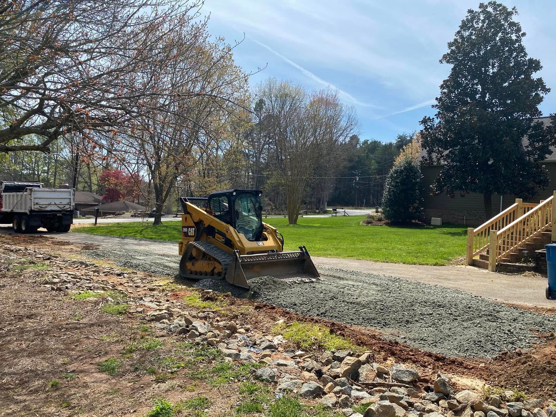 Yellow skid steer leveling gravel in a yard beside a house and wooden stairs