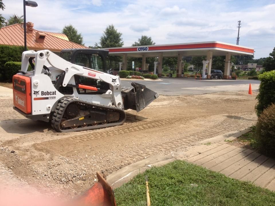 Skid-steer loader spreads gravel near a gas station entrance on a cloudy day