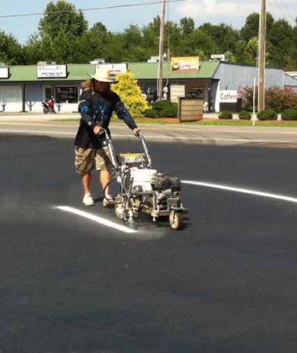 Person operating a line-marking machine on a paved parking lot with store fronts in the background