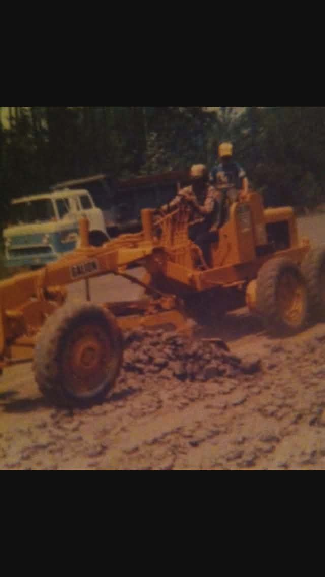 Orange road grader with two workers on a dirt road, surrounded by trees and a truck in the background