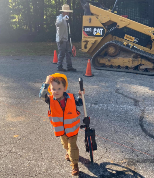 Child in an orange safety vest carrying a toy tool at a roadside construction site with cones and a CAT loader.