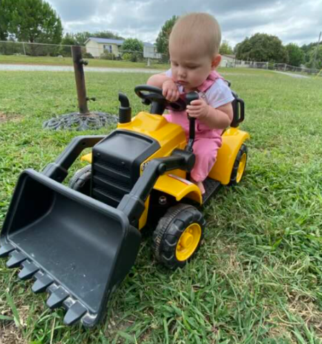 Toddler riding a yellow toy bulldozer in a grassy park, looking down at the controls.