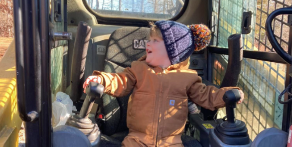 Child in a brown coat and patterned hat sitting in a vehicle cab, holding the controls with gloved hands.