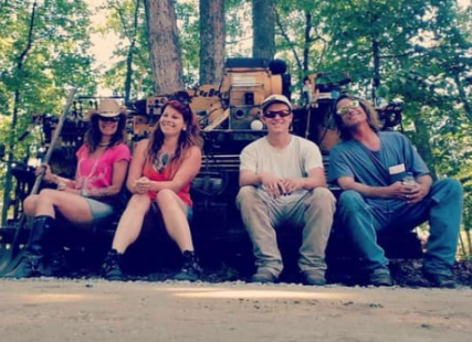 Four friends sitting on a bench outdoors in a wooded area, smiling and relaxed.