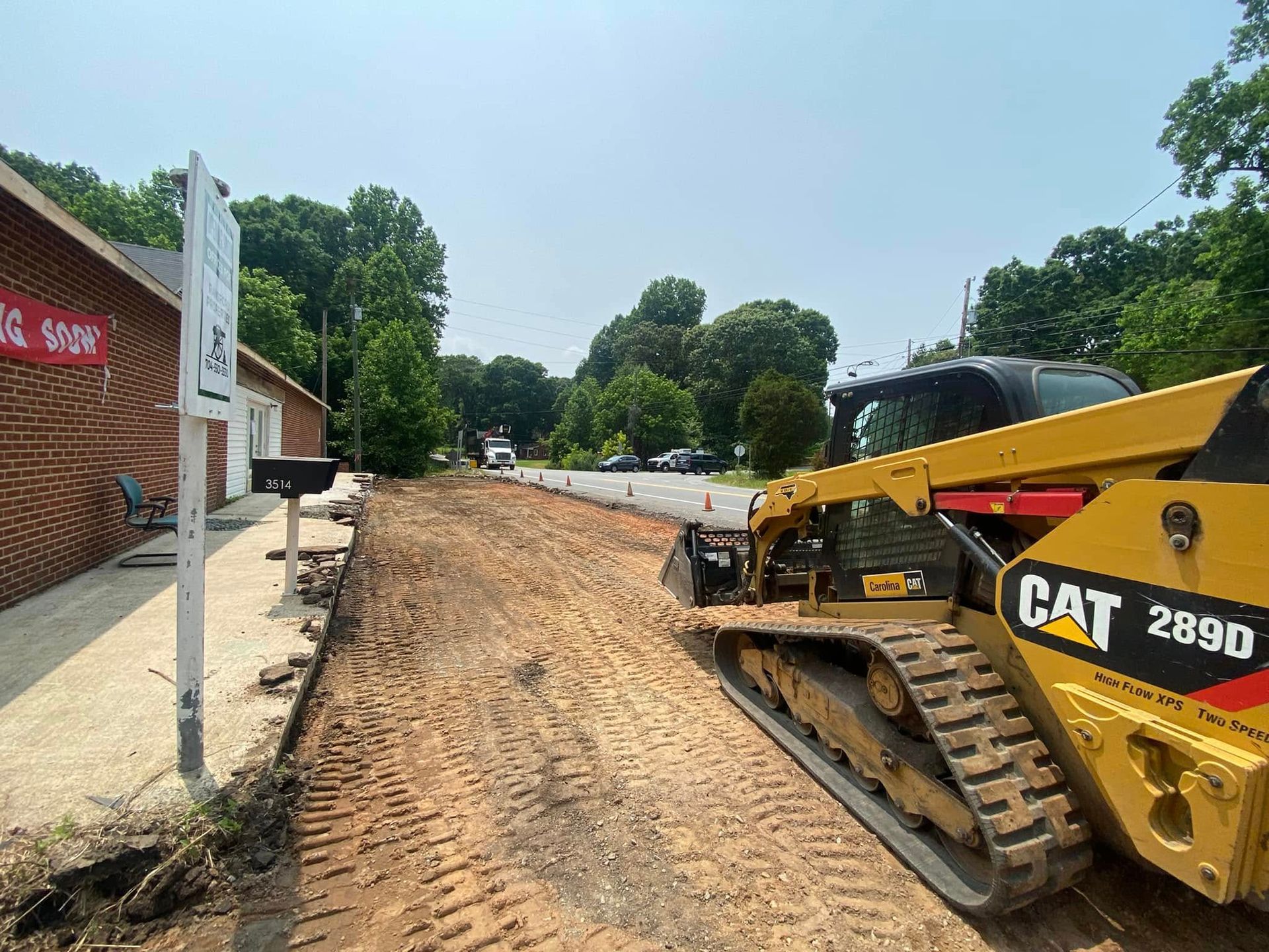 Construction excavator grading a dirt roadside beside a brick building on a sunny day