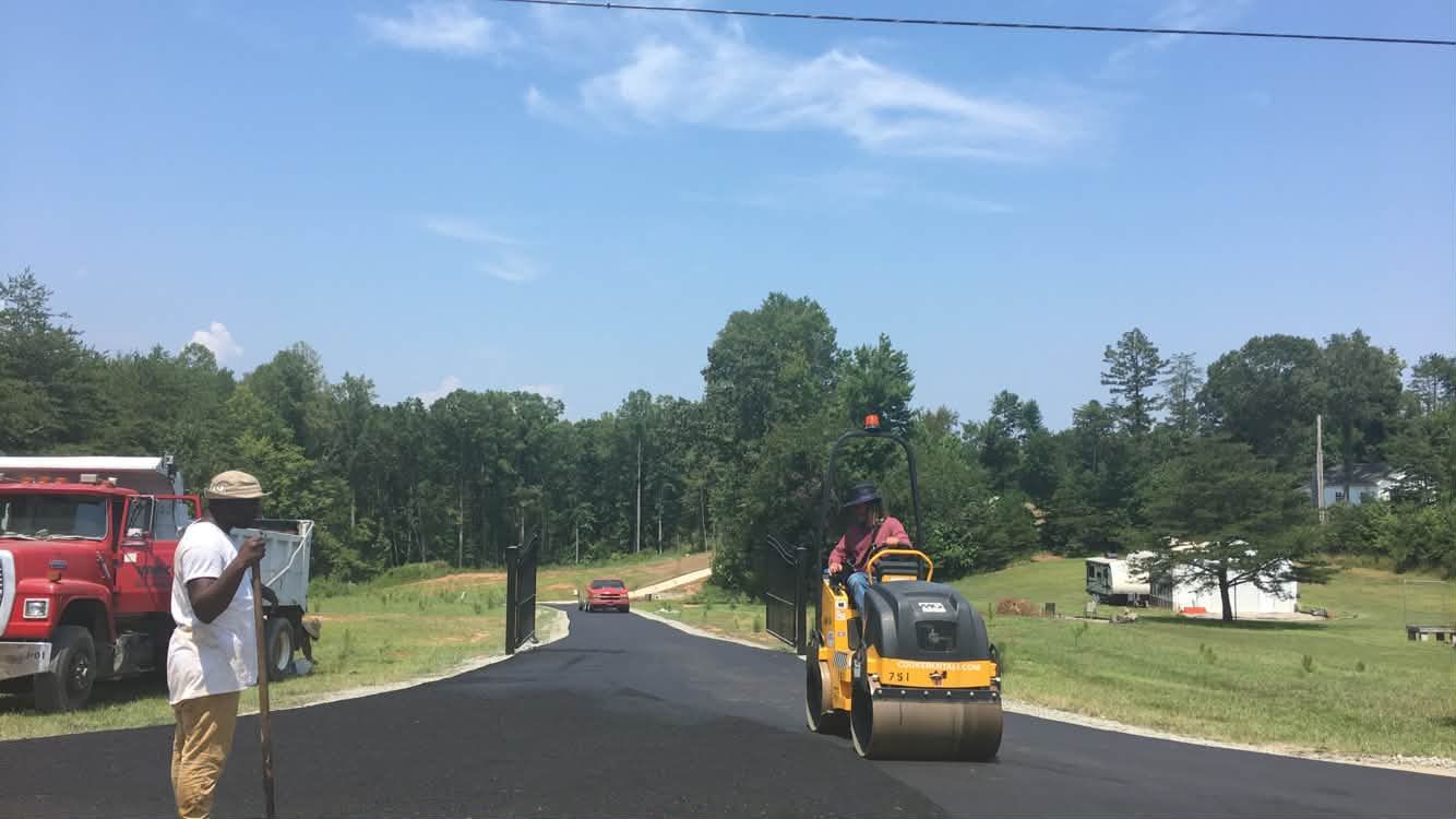 Road workers paving a rural road, with a yellow roller and a red truck nearby.