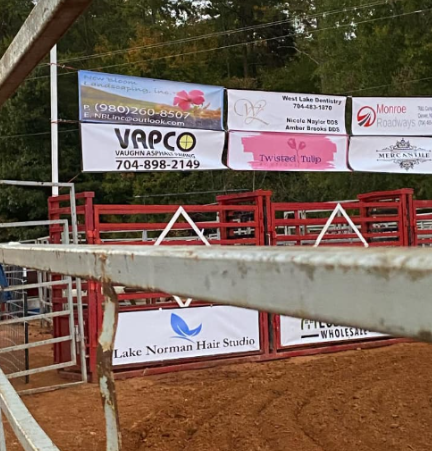 Rodeo arena fence with sponsor banners and a red gate in front of a dirt ring