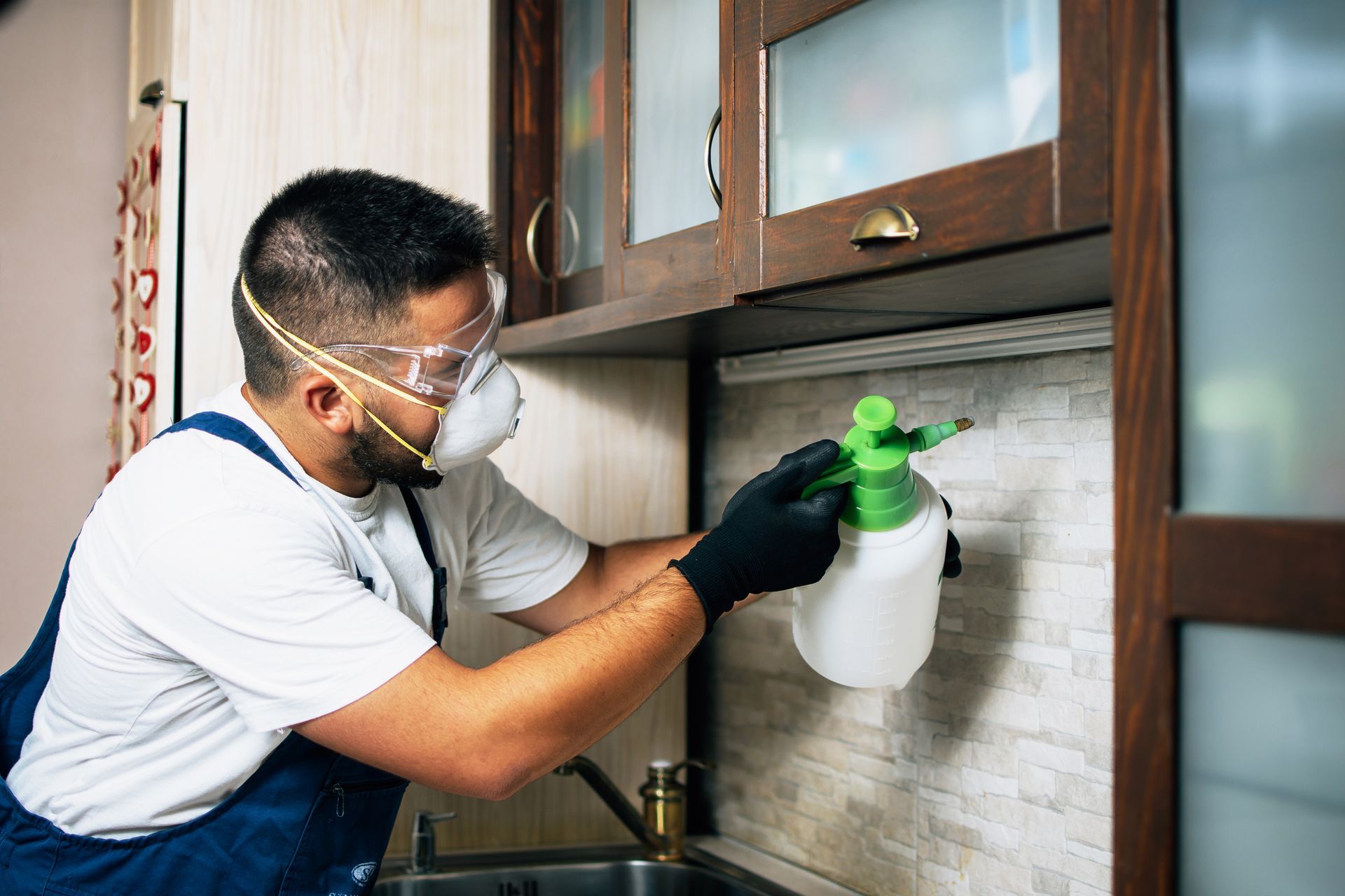 Technician in mask spraying kitchen cabinets during indoor pest control service for insect removal.