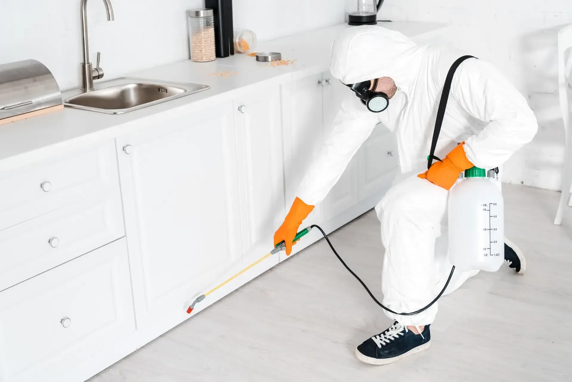 Pest control worker in protective suit sprays base of white kitchen cabinets.
