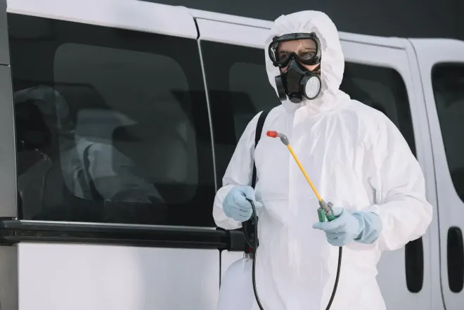 Person in hazmat suit with respirator spraying near a white van.