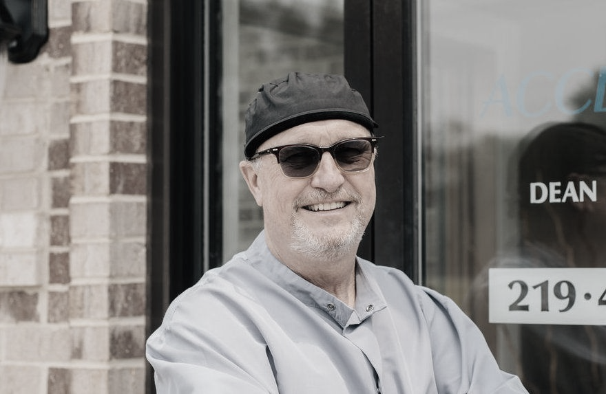 Man in sunglasses and surgical cap smiles in front of a glass door.