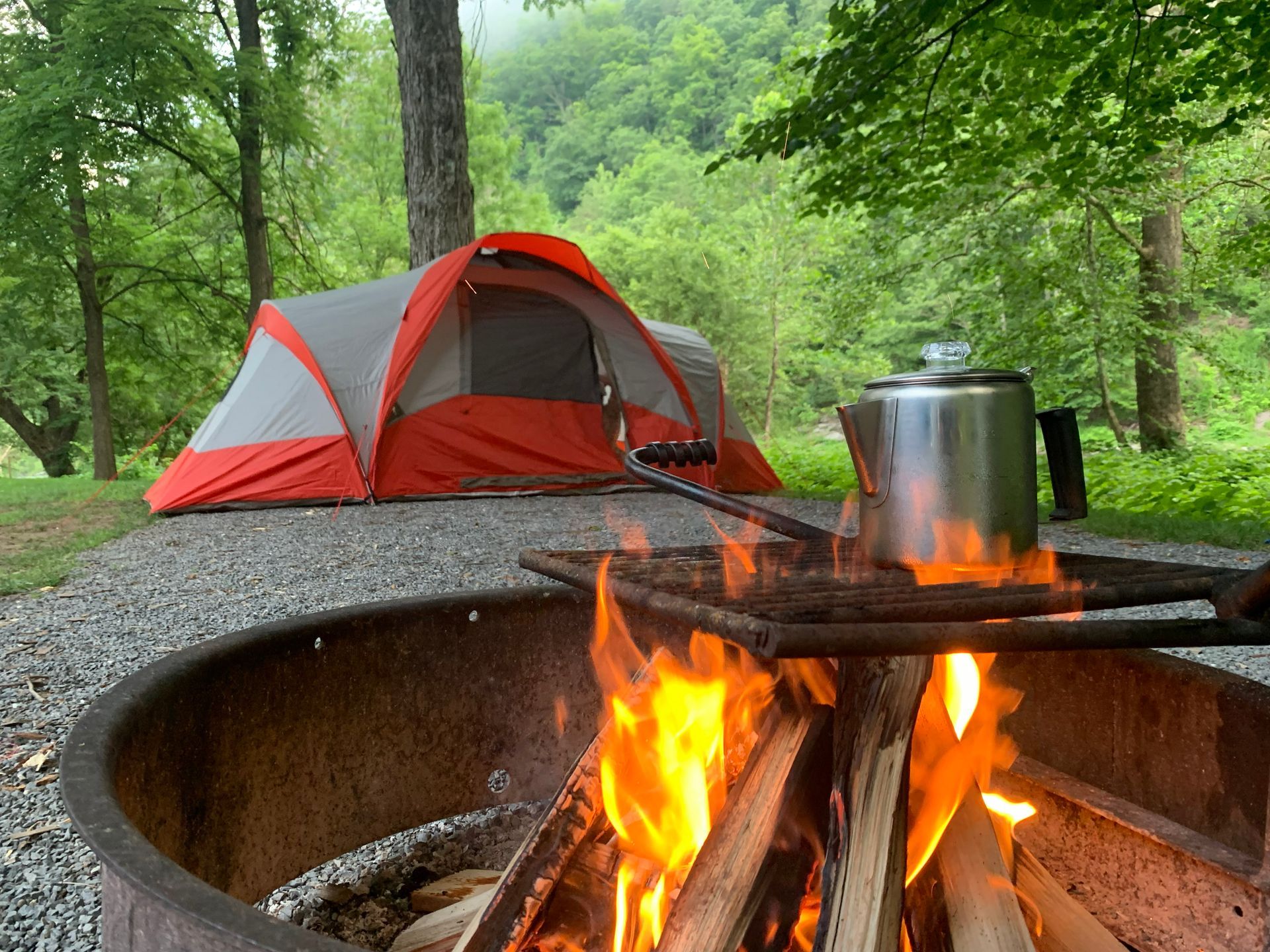 Orange tent near a campfire with coffee pot. Trees and greenery in the background.