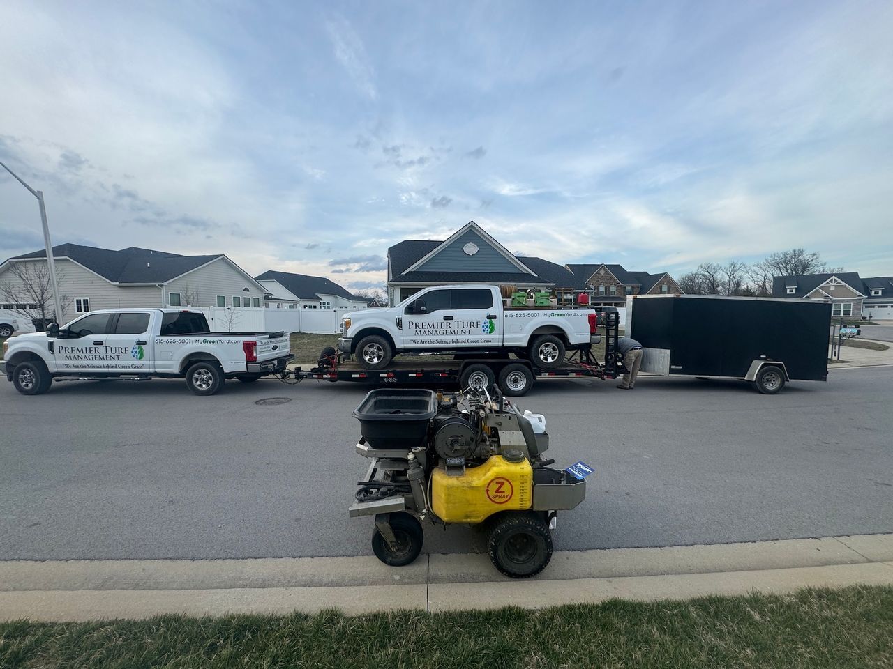 Yellow riding mower in the foreground with work trucks and a trailer parked on a residential street.