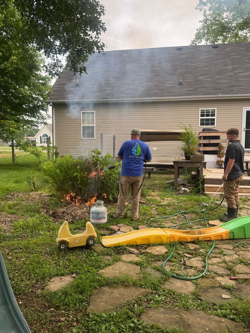 Two people using a hose near a beige house, with smoke or mist rising from the yard.