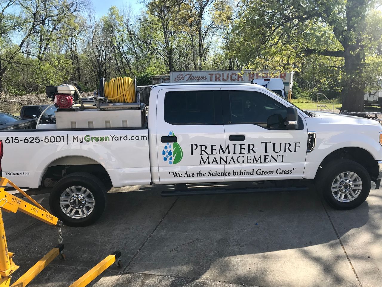 White Premier Turf Management pickup truck with company logo parked outdoors among trees.