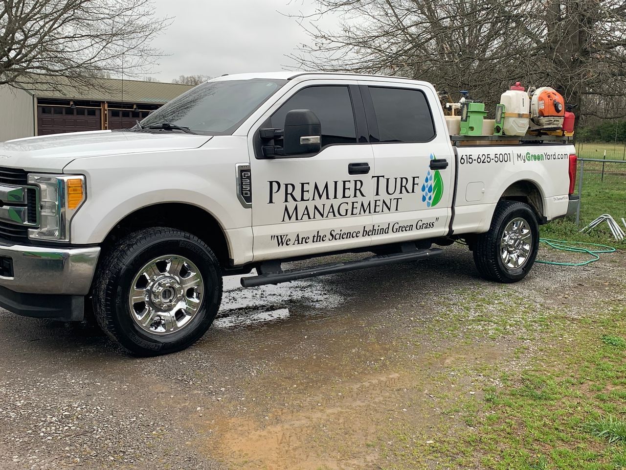 White pickup truck with Premier Turf Management logo parked on a muddy driveway, carrying landscaping equipment.