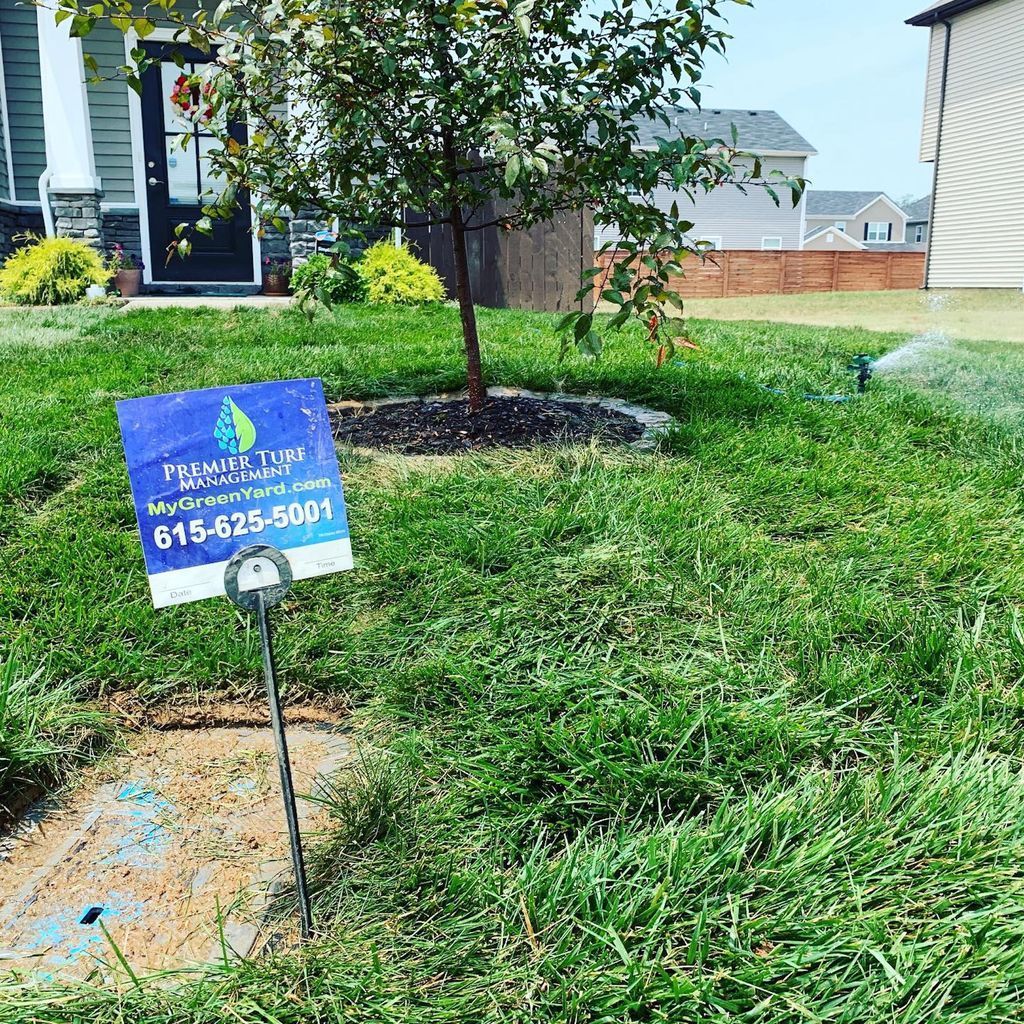 Blue lawn sign on a grass yard with a young tree and suburban houses in the background.