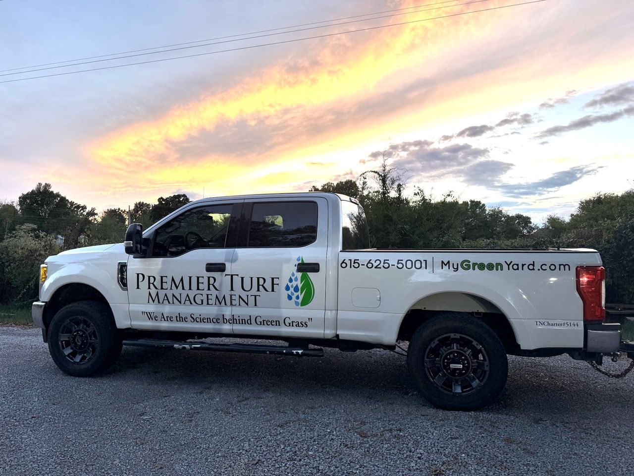 White turf management pickup truck parked on gravel at sunset with a pink-and-gold sky.