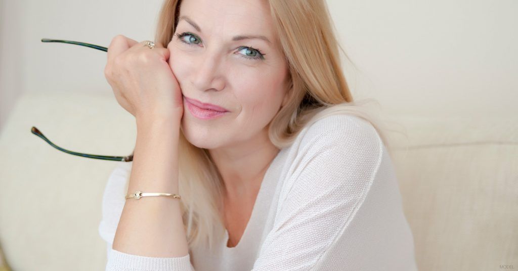 Blonde woman resting chin on hand, holding glasses. Smiling, wearing white, sitting inside.