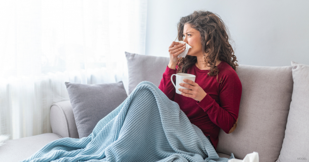 Woman on a sofa covered in a blue blanket, holding a mug and tissue, blowing her nose.
