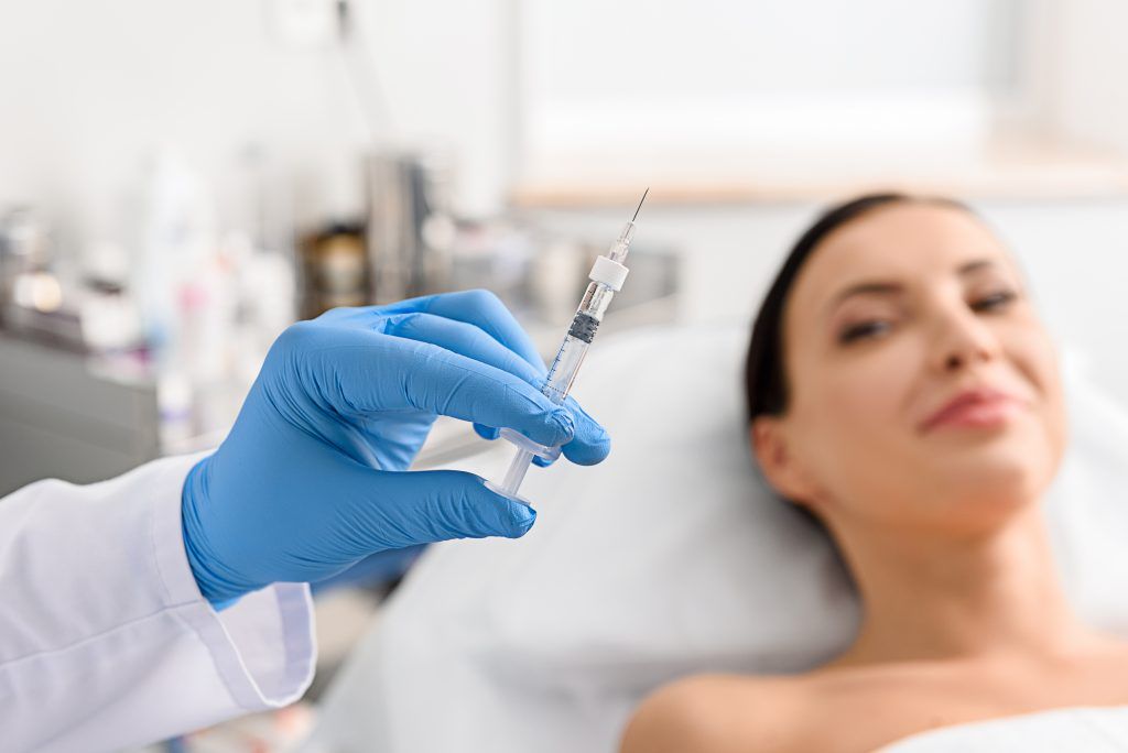 Gloved hand holding syringe near a woman's face, possibly for a cosmetic procedure, in a medical setting.