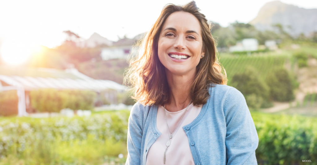 Woman smiles, wearing blue sweater, outdoors in front of greenery; sunlight in background.