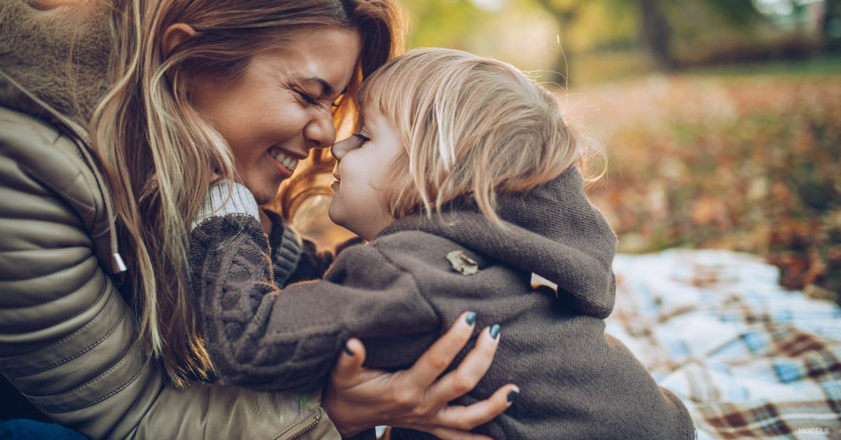 Woman and child embrace outdoors; both smiling, autumn leaves in background.
