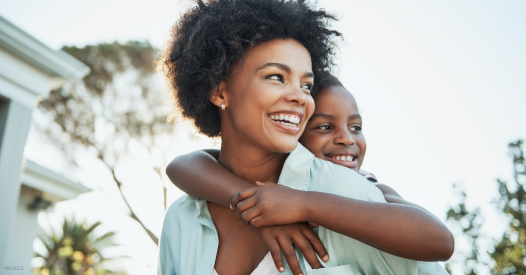 Woman smiles while carrying a child on her back outdoors.