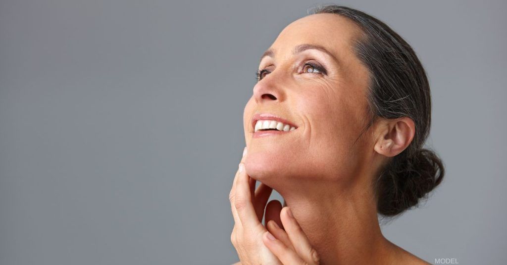 Woman smiling, looking upward, touching her neck, against a gray background.