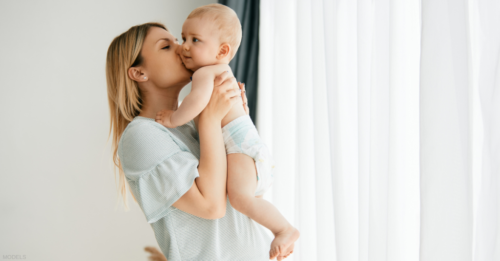 Woman holding baby, kissing its cheek near a window.