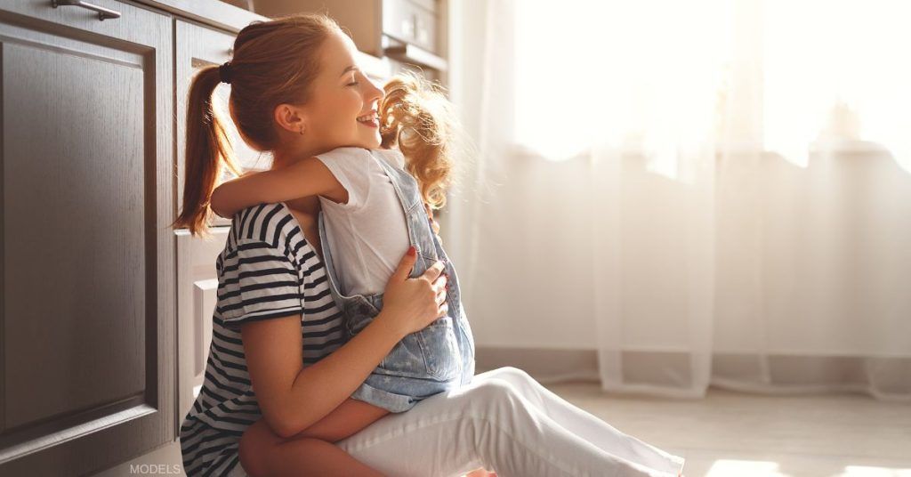 Woman with a ponytail embraces a small child in a sunlit room, both smiling.