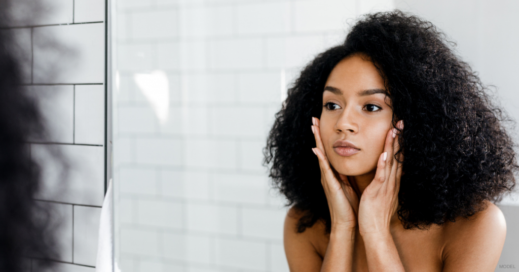Woman looking at her reflection in a bathroom mirror, touching her face. White tiled wall background.