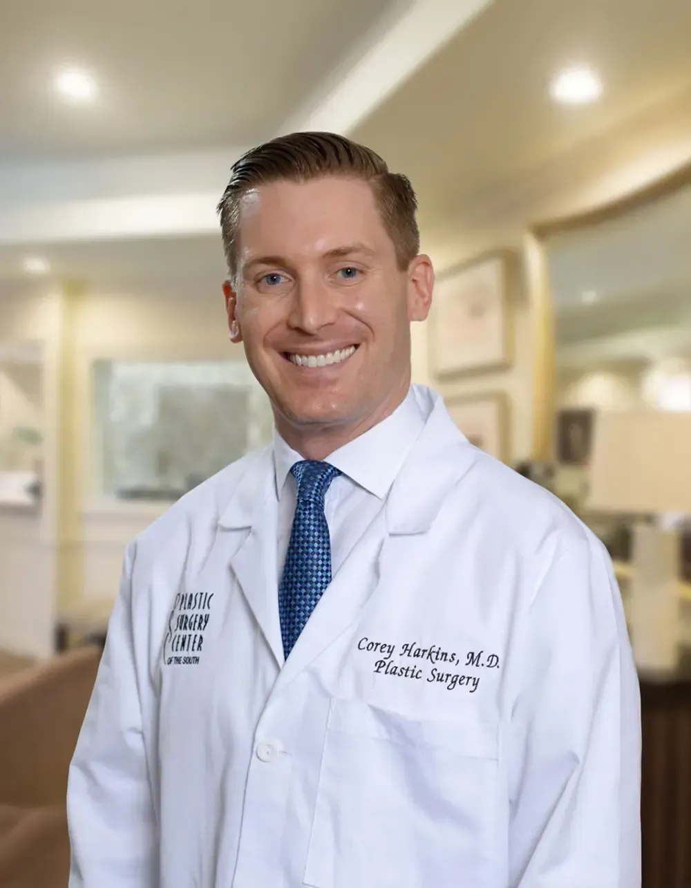 Man in white coat, smiling, tie, standing in what appears to be a medical office.