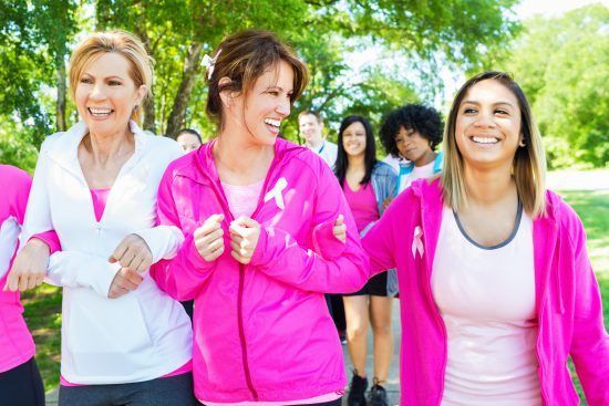 People in pink outfits walking outdoors, arm-in-arm, smiling, with others in the background, possibly at a breast cancer awareness event.