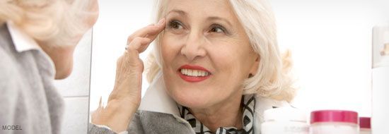Woman looking in a mirror, touching her face, and smiling. There is a jar of cream in the background.