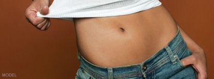 Person lifting a white shirt to expose their navel; wearing blue jeans. Brown background.